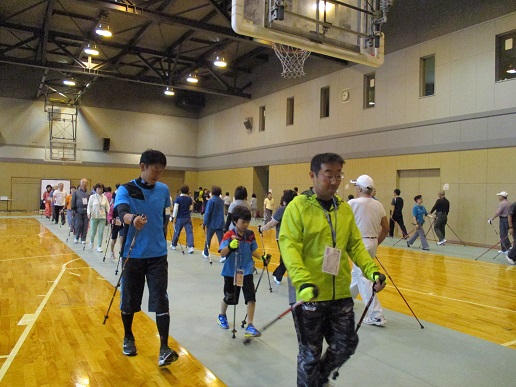 参加者が阿部講師から歩き方の指導を受けながらノルディックウォーキングを行なう様子の写真。